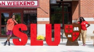 Bold, red styrofoam letters spell S L U. Two students stand on either side of the letters outside of a modern brick building.