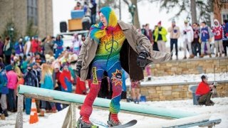 A photo of students at rail jam in brightly colored clothes. In the foreground is a person in rainbow clothes skiing on the rail as everyone behind him watches.