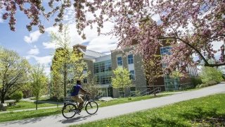 A student riding a bike on a sunny spring day on campus with a stone building in the background