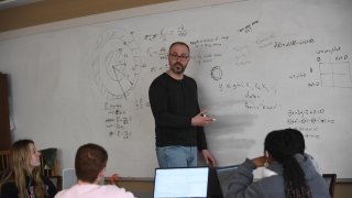 Professor Ivan Ramler stands at the front of a class explaining an equation on a whiteboard. 