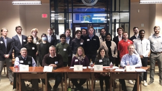 A large group of Saint Lawrence University students stand behind a table of alumni judges.