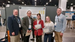 Two female college students hold award certificates and stand in front of their research poster at a geology conference in a large hall. Their professor and two judges stand next to them.