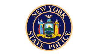 The New York State Police logo is a blue circle with two tan bands, one on the exterior edge, the other just inside. Text that follows the circle on the inside the outer band reads "NEW YORK STATE POLICE" ... inside the inner-most circle is the New York State Coat of Arms, the center of which shows a ship and sloop on a river bordered by a grassy shore and a mountain range with the sun rising behind it. Liberty and Justice stand on either side, under an American eagle.