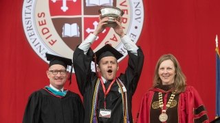 Michael Ranger and Kathryn Morris, wearing commencement regalia, smile as Charlie Reinhardt holds a silver bowl above his head in celebration. A scarlet banner with the Saint Lawrence crest is in the background.