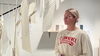 Photo of Saint Lawrence Female Student wearing a white Saint Lawrence University Crew Neck, staring intently at the three dimensional art piece pertruding from the wall. The photo is taken through the art of her. The artwork is a closeline style display of papers with words hanging.