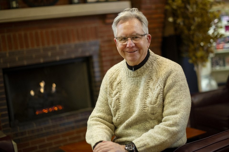 A photo of Bill Martin sitting and smiling in front of a fireplace.