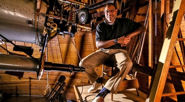 Derrik Pitts sits on a chair inside of an observatory with wood walls. A very large telescope is in front of him. 