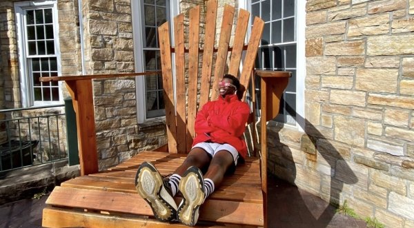 A person, wearing a scarlet jacket and eclipse sunglasses, sits on a giant Adirondack chair and smiles up at the sun.