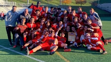 The Saint Lawrence Men's Soccer Team, wearing scarlet soccer jerseys, pile together on the field to celebrate winning the Liberty League Championships.