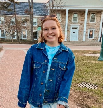 Teal Borden, class of twenty-twenty-two, wearing a jean jacket and blue dress and walking on a pathway on a college campus.