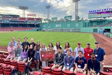 A group of students holding a Saint Lawrence University flag stand in the stands in front of the field at Fenway Park.
