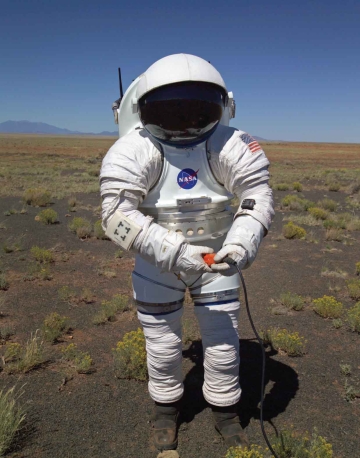Dean Eppler, wearing a white NASA suit, tests a bright orange button in the desert.