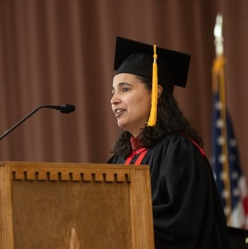 Ana Estevez, wearing black commencement regalia, stands at a podium and addresses the crowd.
