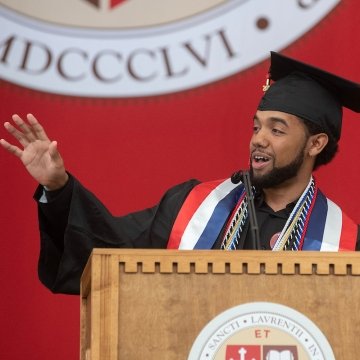 Brian Uceta, wearing commencement regalia, stands at a podium and addresses the Class of 2023. A scarlet banner with the Saint Lawrence logo hangs in the background.