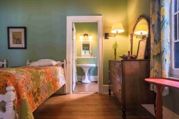Two white single beds with orange floral quilts in a light green bedroom. There is a dark brown wooden dresser next to a doorway that leads to a bathroom that features a white porcelain sink.