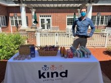 St Lawrence alumni Conor McDermott standing at a table full of snacks and drinks outside the brick wall entrance of Kind Senior Care, a nonmedical home care business.