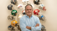 Brian McCarthy folds his arms and stands in front of a display of National Football League helmets.