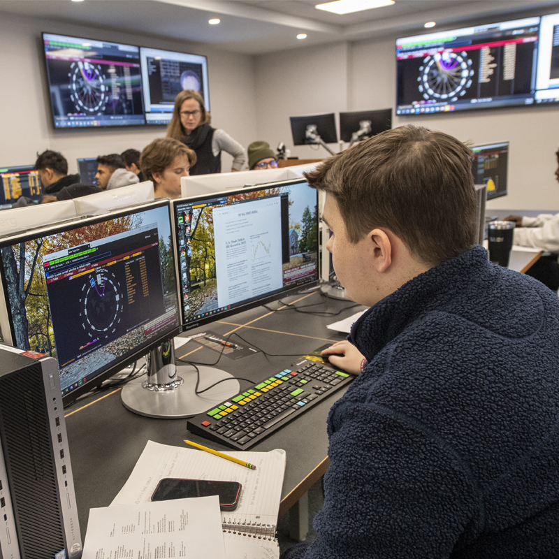 A Saint Lawrence student analyzes data on a computer monitor in the Bloomberg Finance Lab. A professor works closely with a student in the background.