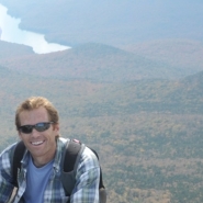 man kneeling on rock with mountain and stream in background