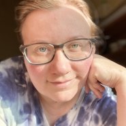 Professor with blonde hair, dark glasses, smiling, leaning on a hand