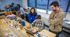 Students collaborate at a workbench in a Saint Lawrence University lab, using a laptop and electronic components. One student types while others stand nearby, observing and discussing the project. Tools, wires, and equipment are spread across the table, showing hands-on learning in progress.