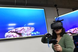A student wearing a virtual reality headset experiments with virtual reality software. There are two large television screens behind them.