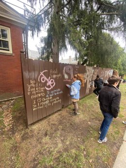 Sara Ashpole stands outside on a sunny day and draws on a brown fence with chalk.