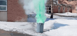 Green mist pours out of a grey garbage can sitting in the snow on a sunny day. 