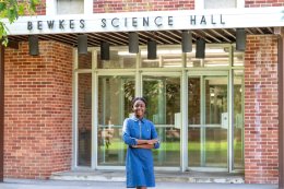 Francesca Mnenula wearing a blue jean dress and standing in front of a mid-century modern brick science building.