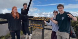 Four smiling students pose and celebrate in front of a sign at Irazú Volcano National Park in Costa Rica, with one student mid-jump. The sign shows an elevation of 3,432 meters above sea level. The sky is partly cloudy with scenic views in the background.