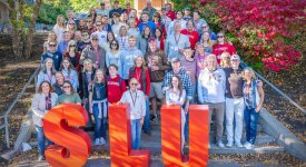 A group of Saint Lawrence alums and current students stand in front of the student center on a sunny fall day. There are large SLU letters in front of the group.