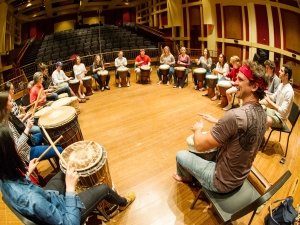 A group of students sit in a drum circle in a large auditorium.
