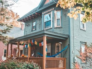 A large teal house with a string of small, yellow, blue, and red flags hanging on the porch. A student sits in a hammock on the front porch.