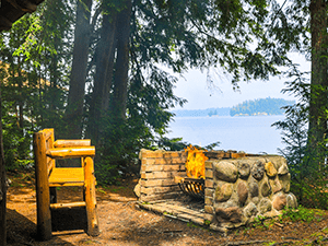 An Adirondack style bench sits in front of a roaring outdoor fire overlooking Upper Saranac Lake.