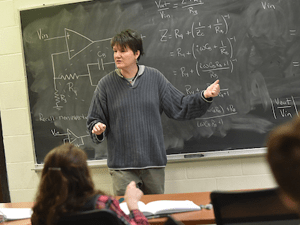 Catherine Jahnke, wearing a long sleeve grey sweater, points at a black chalk board with physics formula. Students look on attentively.