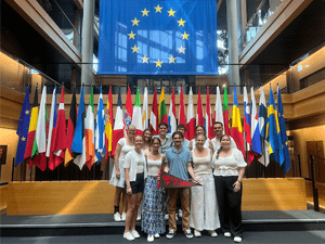 A group of Saint Lawrence students hold a scarlet and brown Saint Lawrence flag while standing in front of several flags at the European Union.