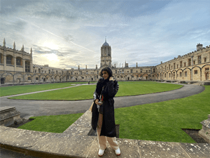 A person wearing a long black trench coat stands in the courtyard of a historic London college.