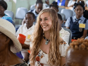 A woman with long dark blonde hair smiles broadly while sitting in a crowd of people.