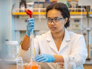 A person wearing safety goggles, gloves, and a lab coat carefully uses a pipette in a laboratory setting. Shelves filled with bottles and equipment are visible in the background. The person appears focused and precise in handling the laboratory tools.