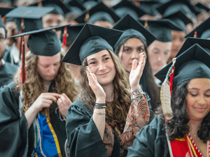 A student in graduation attire smiles and claps during a commencement ceremony, surrounded by classmates in caps and gowns. The group appears joyful and proud, celebrating their achievements. The scene captures a sense of accomplishment and unity.