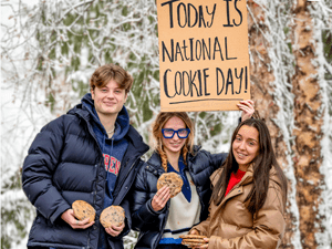 Three people stand outside in winter clothing, holding large cookies, with one person holding a sign that reads, &quot;Today is National Cookie Day!&quot; Snow-covered trees are visible in the background.