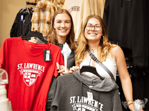 Two students smile while holding up Saint Lawrence University apparel in a campus store. One holds a red T-shirt, and the other displays a gray sweatshirt, both featuring the university’s crest. The scene captures school spirit and enthusiasm.