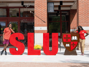 Two students, wearing scarlet shirts, stand on each end of large S, L, U letters, and the shield, in front of the Sullivan Student Center on the Saint Lawrence University campus.