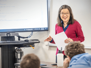 Grace Huang, wearing a red sweater, stands in front of a classroom and smiles at students. There is a large digital screen in the background.