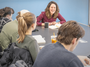 A professor in a maroon top smiles while leading a discussion with students seated around a table. The students, some taking notes and others listening, have books in front of them. The setting is a bright classroom with a blue wall in the background.