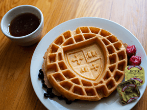 A waffle with a crest design on top, served with blueberries, raspberries, butter packets, and syrup on the side.