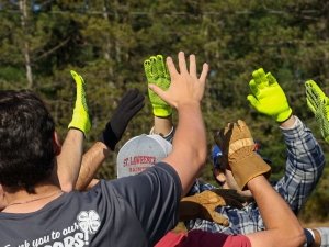 A group of people wearing work gloves join hands in a team huddle outdoors, with one person wearing a Saint Lawrence University cap.