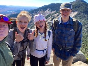 Four hikers smile for a selfie at a mountain summit with a scenic view of forested hills and valleys behind them.