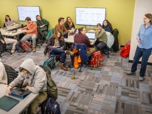 Lisa Torrey stands at a whiteboard pointing to a diagram while students sit at tables in small groups, listening, writing, or working on laptops in a bright, modern classroom.