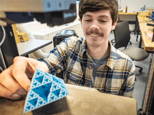 A person placing a blue triangular 3D-printed object into a machine in a workshop setting.